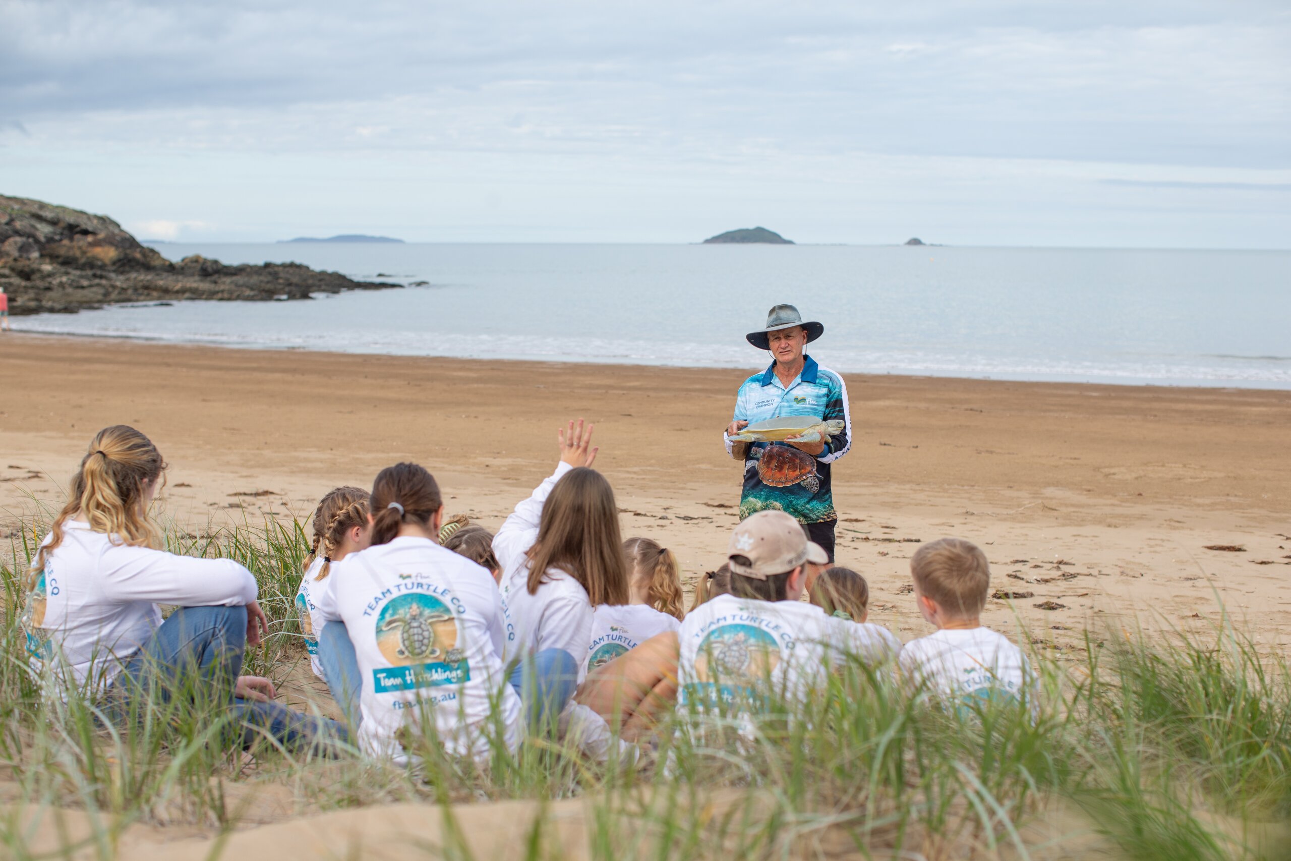 An FBA volunteer shows a model of a turtle to a group of kids on the beach wearing FBA Team Turtle shirts